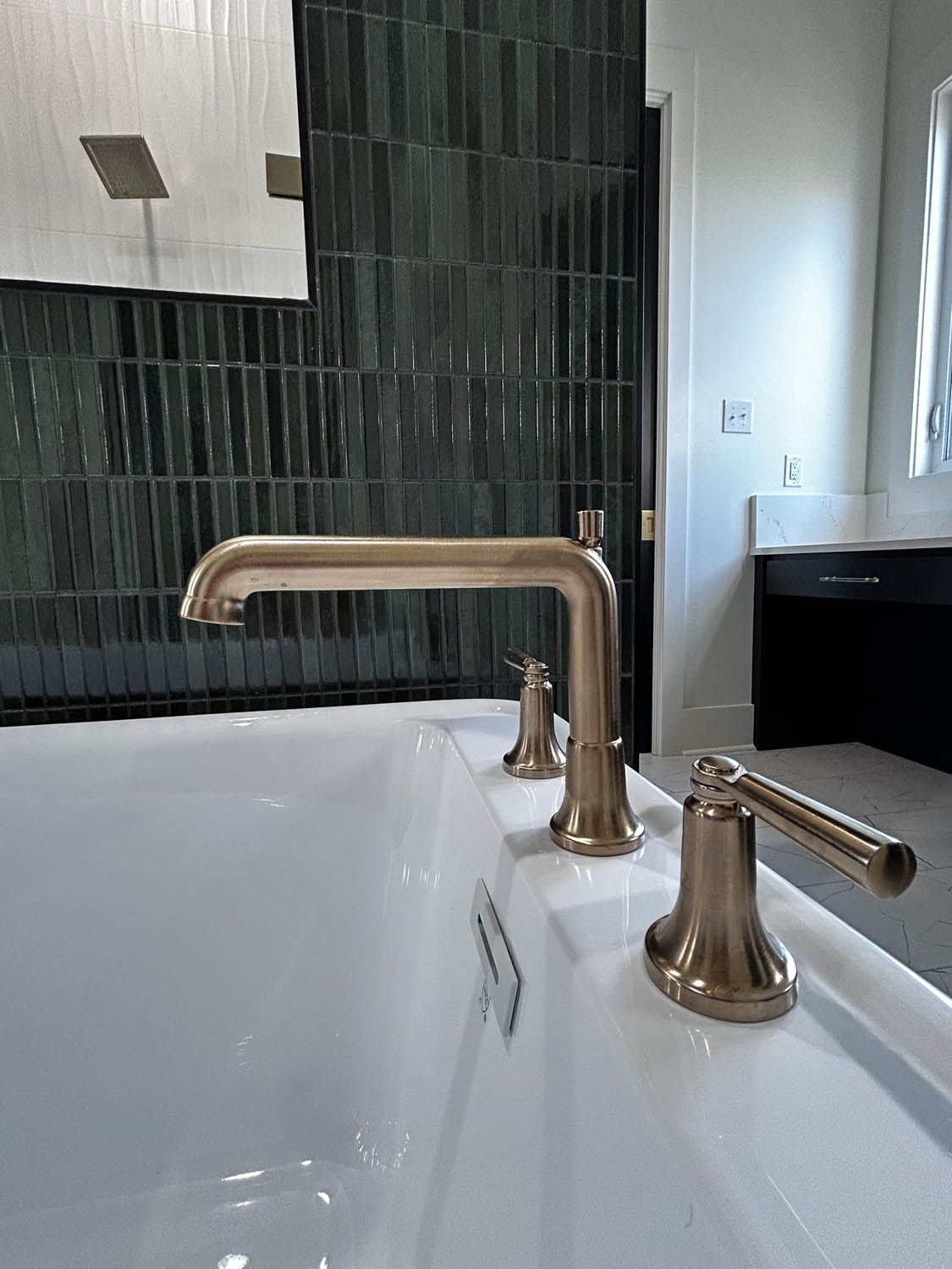 Gold bathtub faucet against a dark green tiled wall. A white tub is in the foreground.
