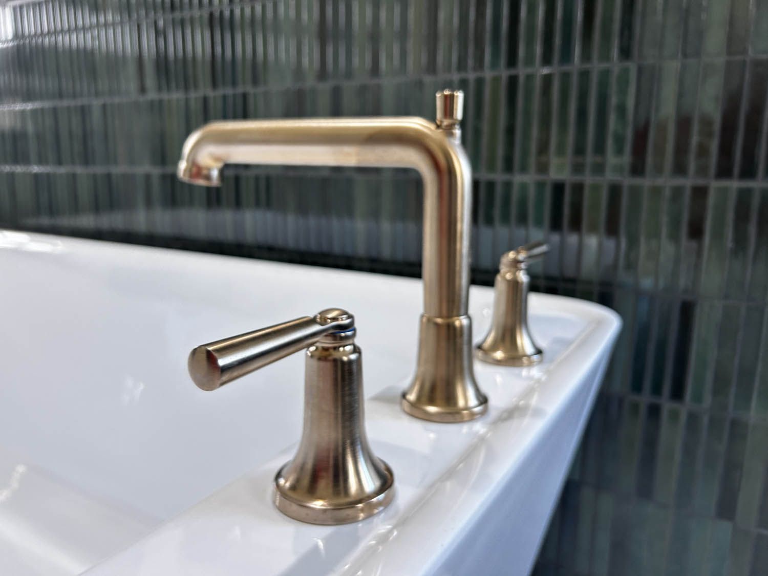 Gold-toned bathroom faucet with two handles and spout, on a white bathtub, against a dark green tiled wall.