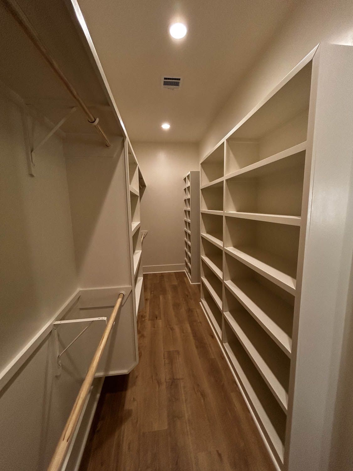 Long hallway with built-in white shelves and a wooden floor. There is a hanging rod and lights.