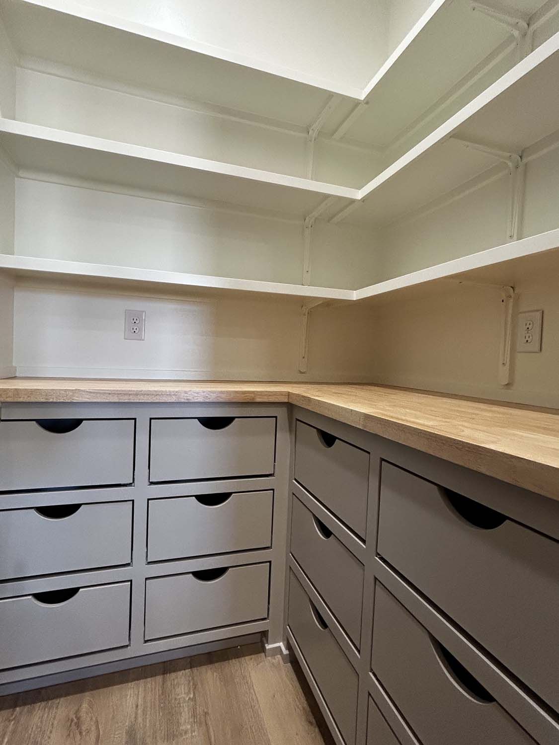 Empty, organized pantry with light gray drawers, wooden countertops, and white shelves.