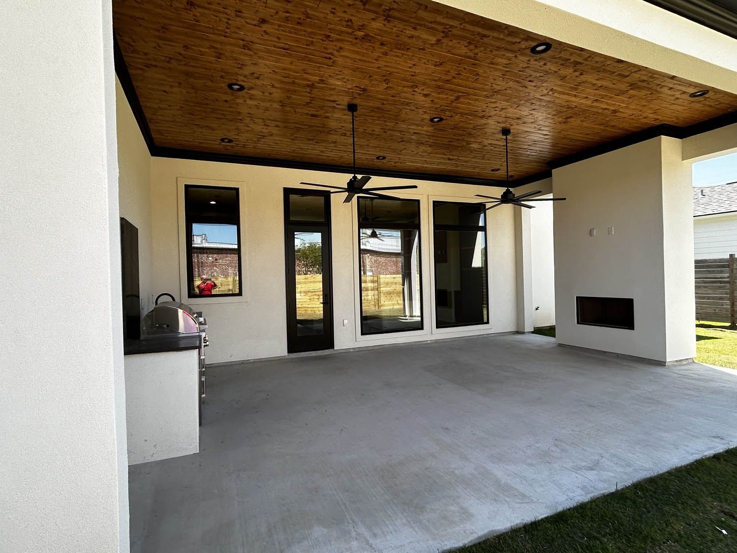 Covered patio with a concrete floor, dark trim, and a fireplace. Ceiling fans hang from a wood-paneled ceiling.