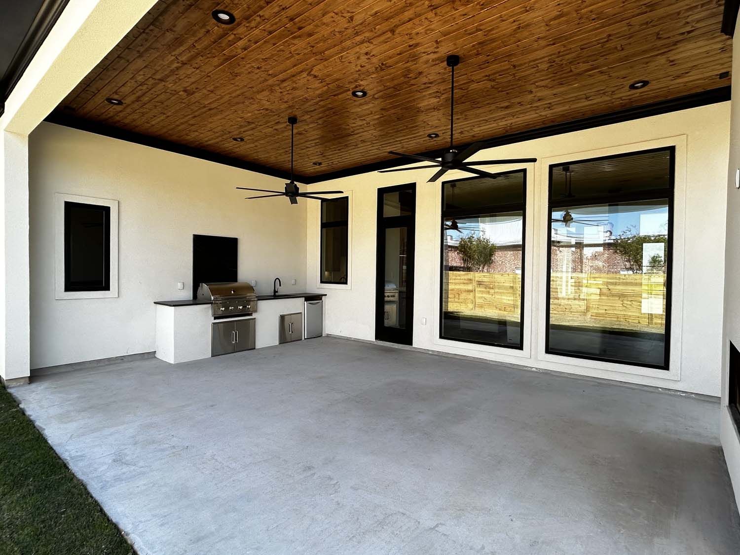 Covered outdoor patio with built-in grill, concrete floor, wooden ceiling, and large windows.
