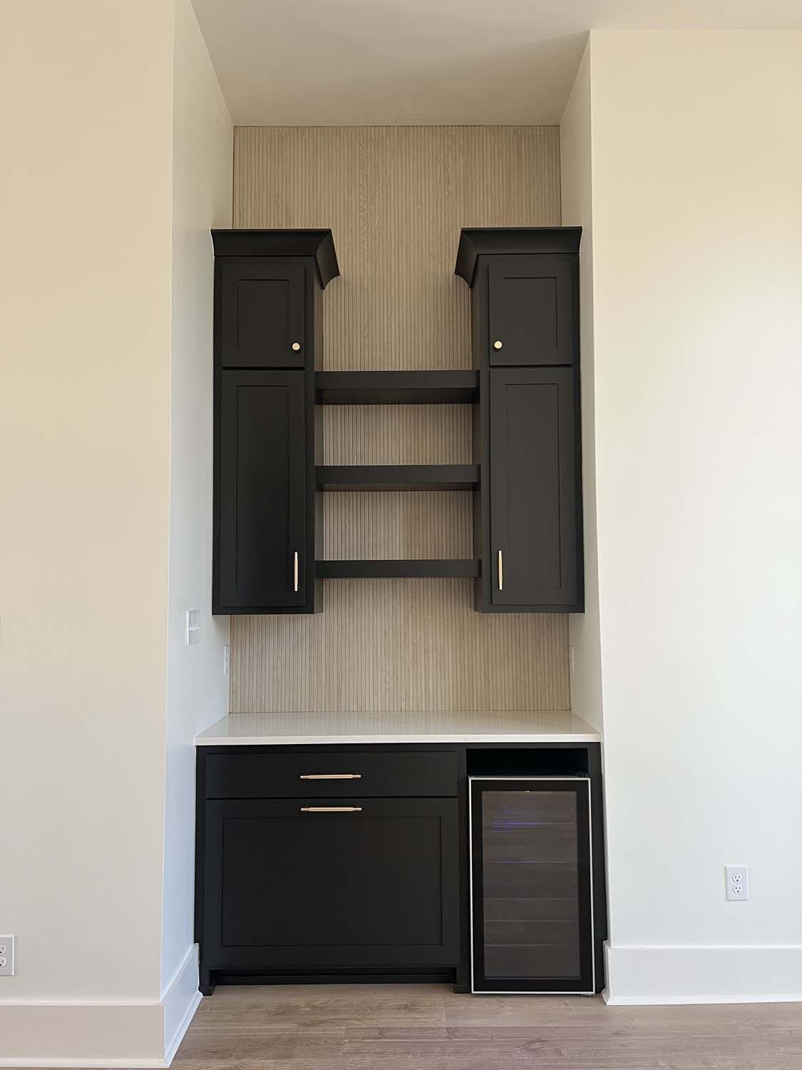 Built-in black cabinetry with open shelves, mini fridge, and countertop against textured neutral wallpaper in a niche.