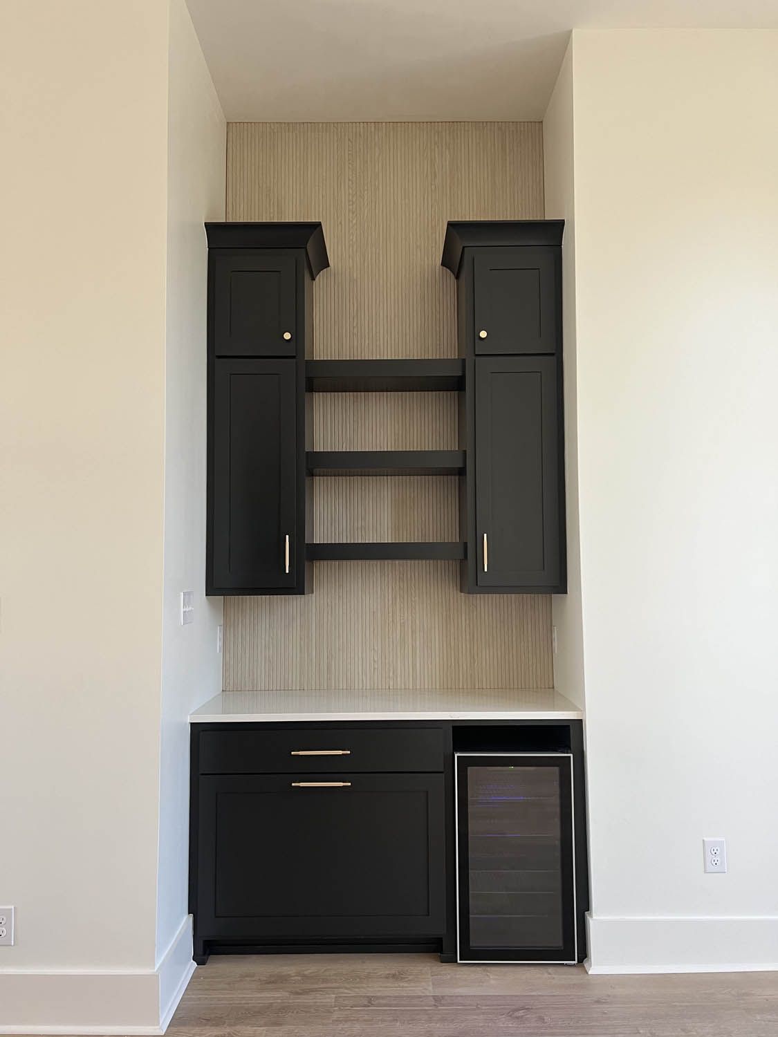 Built-in black cabinet with shelves, countertop, and wine fridge in a recessed wall, with beige wallpaper.