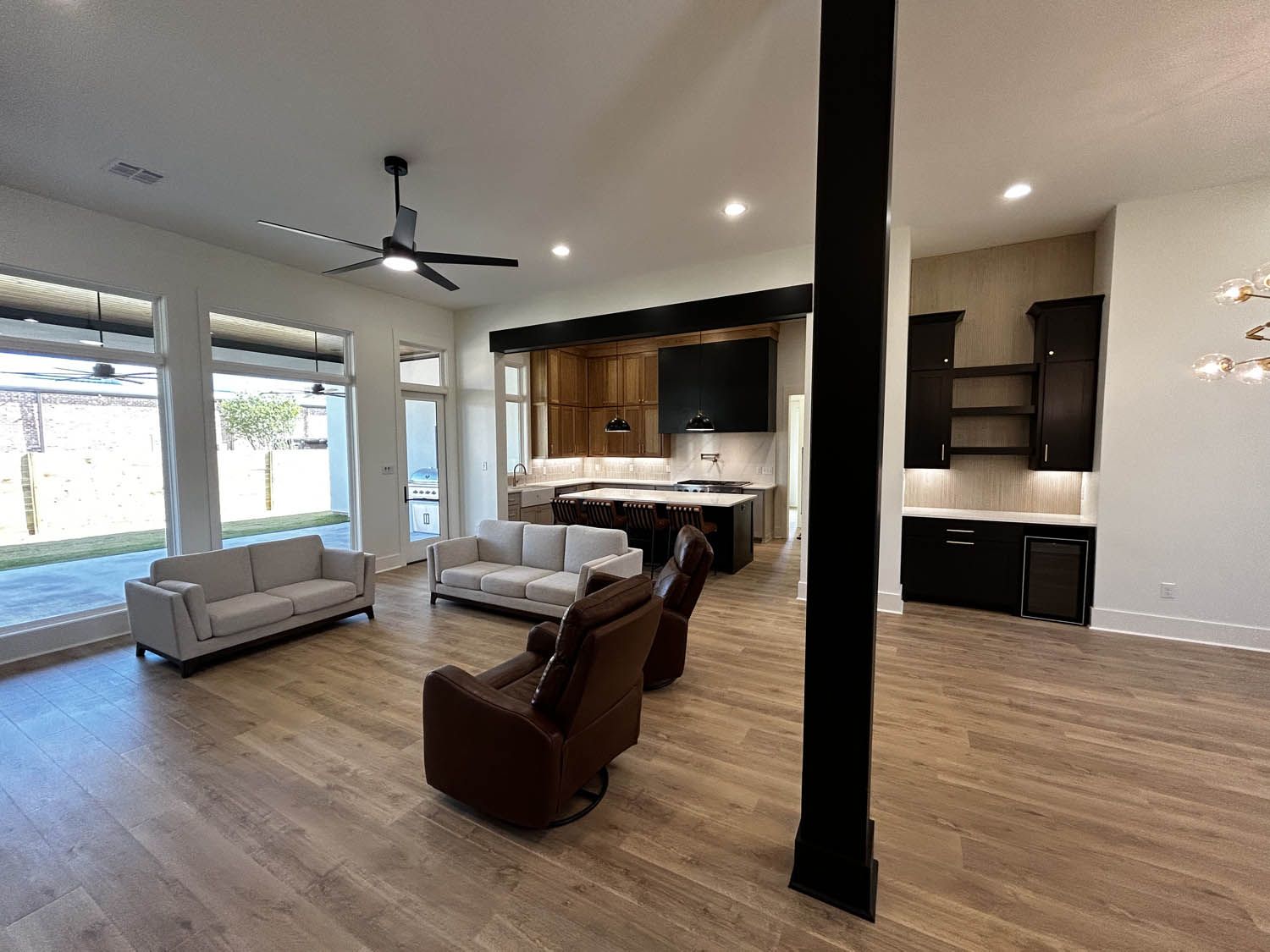 Spacious living area with couches, chairs, and kitchen island. Large windows overlook backyard. Wooden floors, black accents.