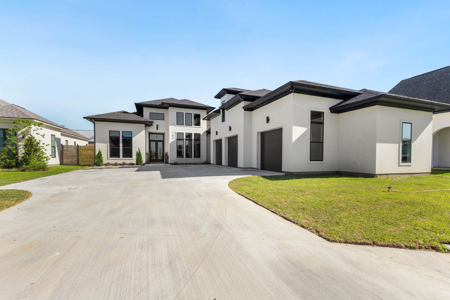Modern, two-story house with stucco exterior, dark roof, and a three-car garage; long concrete driveway and green lawn.