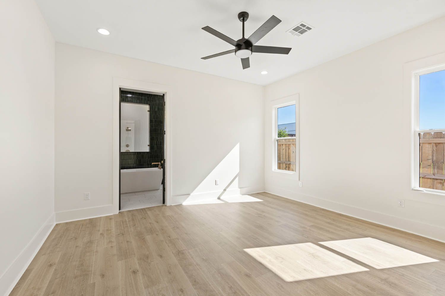 Empty bedroom with white walls, wood floors, and ceiling fan; a bathroom door is visible.