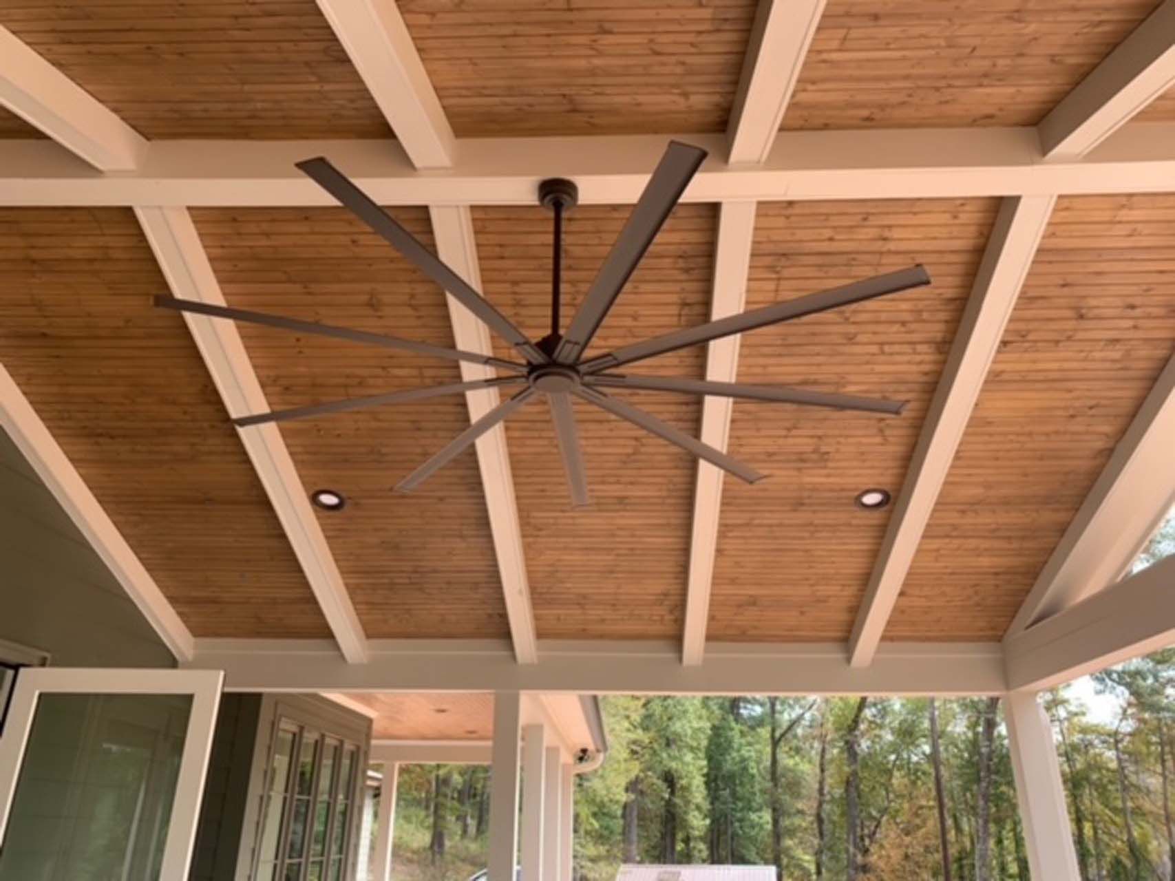 Covered porch ceiling with large ceiling fan, wooden planks, and white beams.