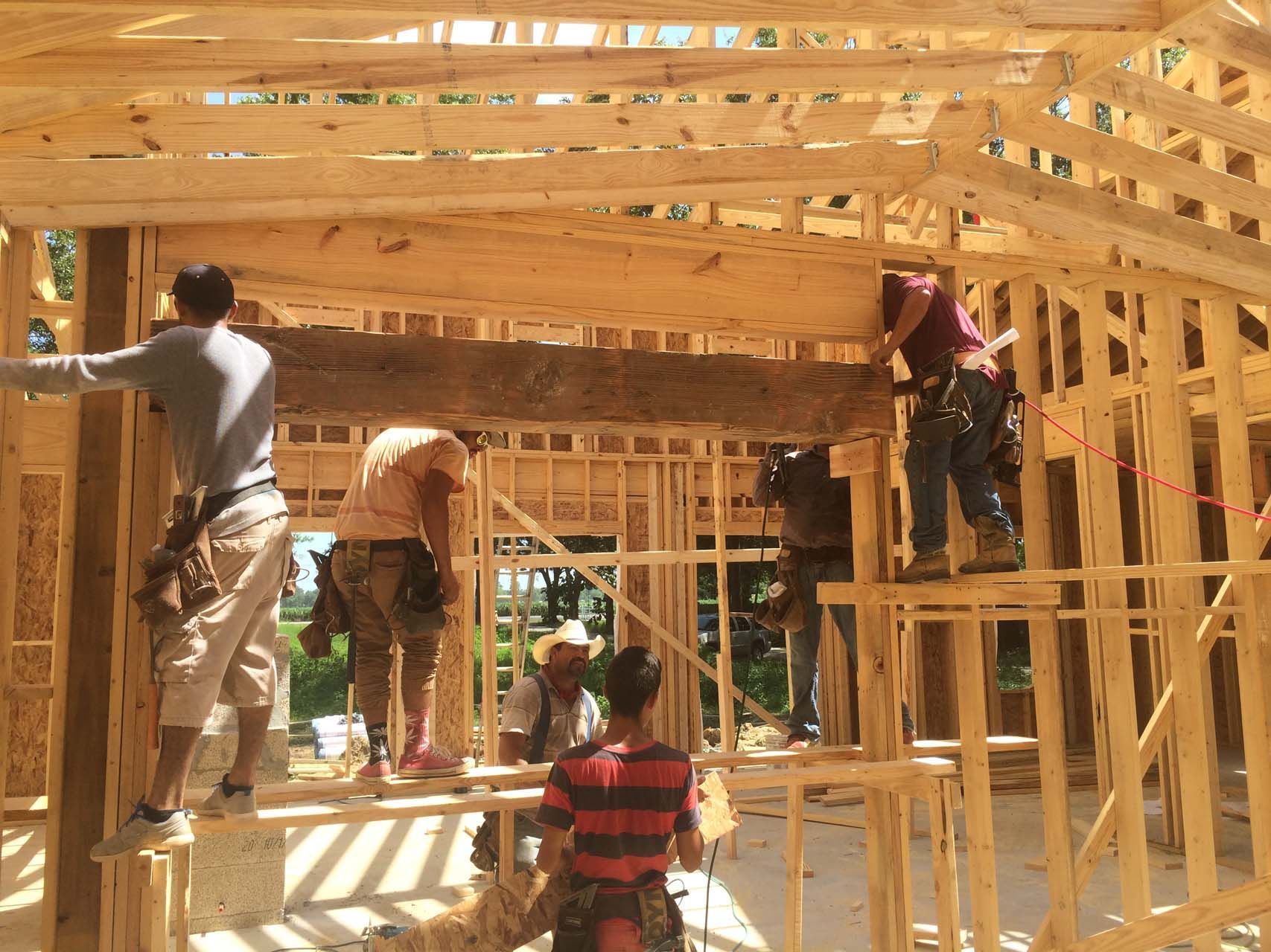 Construction workers framing a wooden structure, installing a large beam. Sunlight streams through.