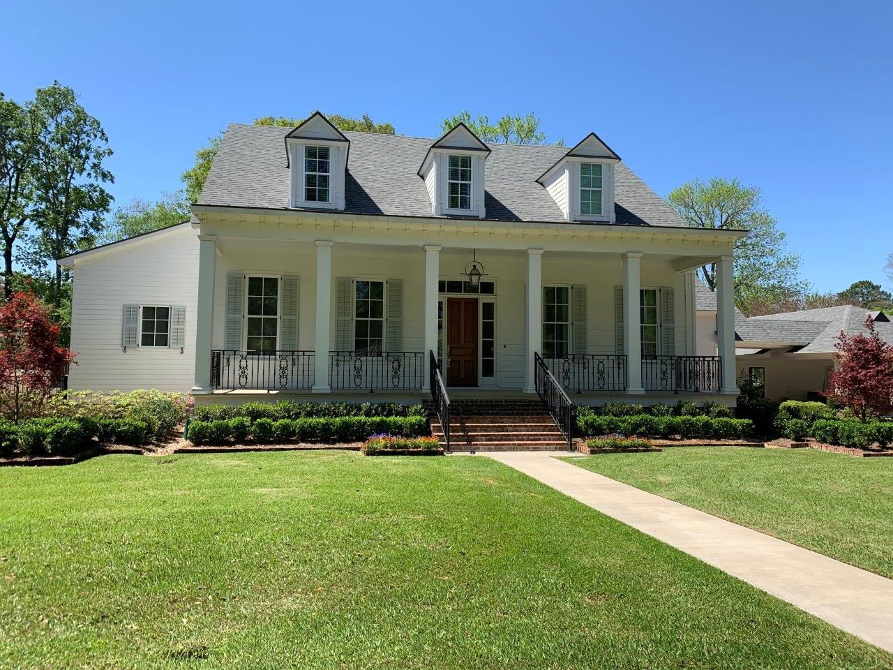 White house with porch, shutters, and dormers. Green lawn, blue sky.