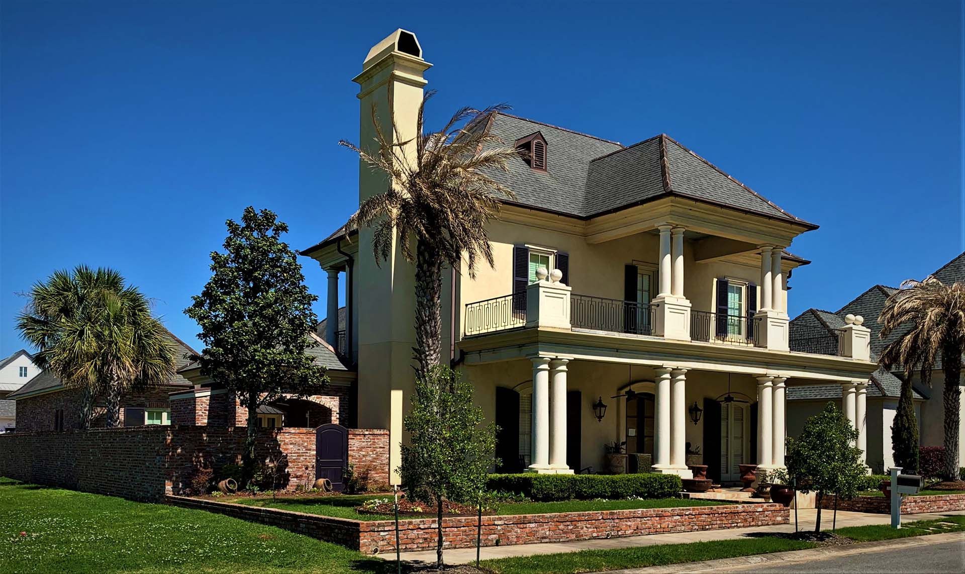 Two-story beige house with columns, balcony, and dark roof under a blue sky, surrounded by greenery.