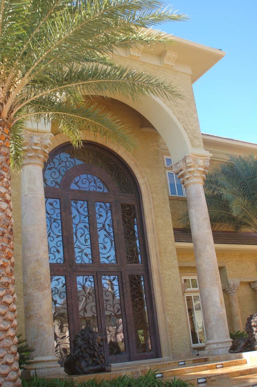 Large house with arched doorway, columns, and ornate wooden door. Palm tree in foreground.