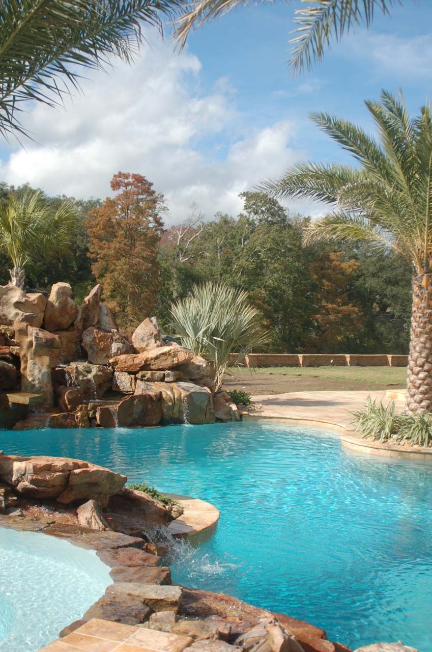 Swimming pool with a waterfall feature surrounded by palm trees and greenery on a sunny day.