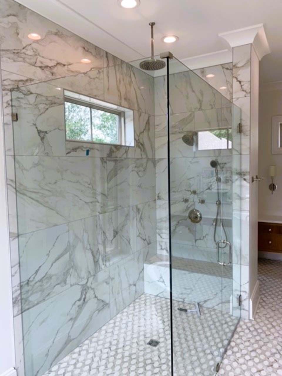 Spacious marble-tiled shower with glass doors and windows; features a rain shower head and white hexagon floor tiles.