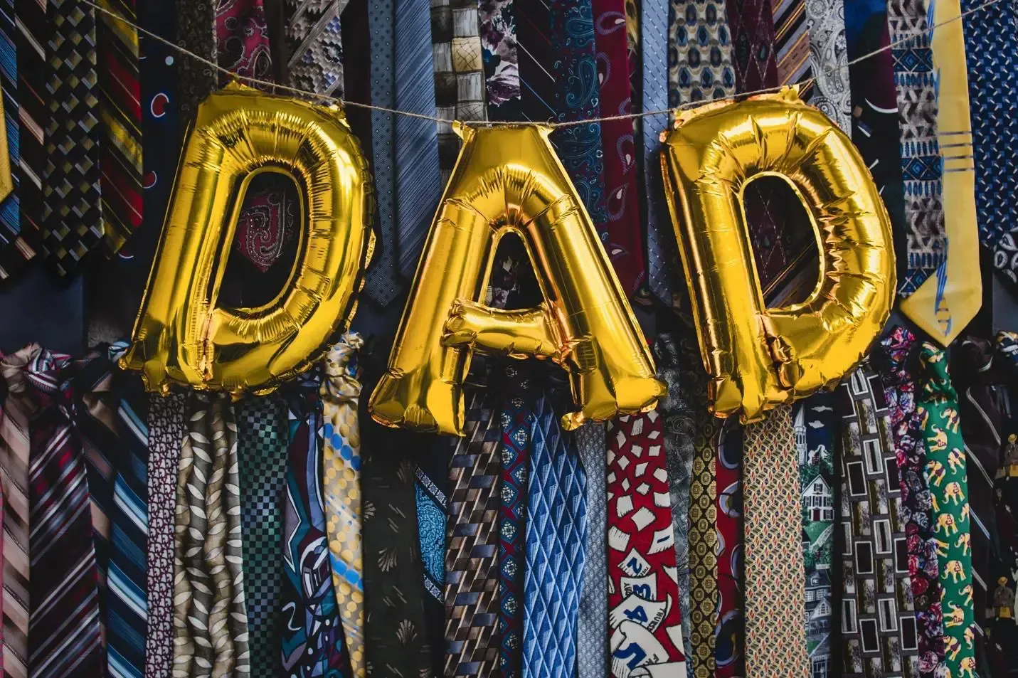 Gold “DAD” balloons hanging over a colorful background of woven neckties