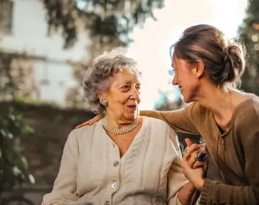 Two women smiling and talking outdoors, one gently touching the other’s shoulder.