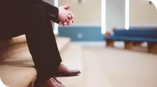 Person sitting on wooden steps with hands clasped, in a bright indoor space.
