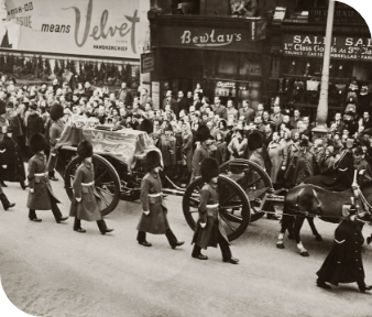 Crowd watches a horse-drawn street parade with uniformed marchers in front of storefronts.