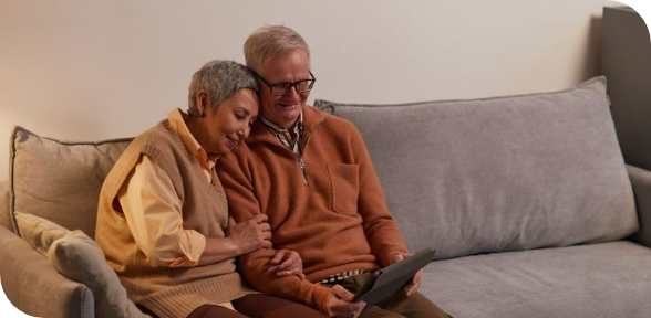 Two older adults sit closely on a couch, looking at a tablet in a cozy living room.