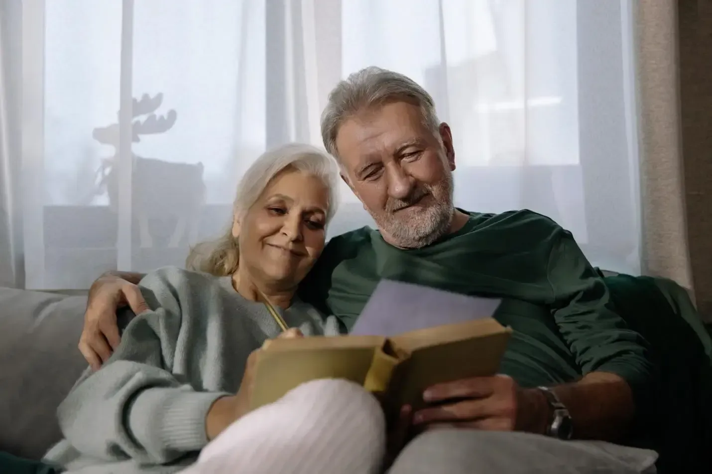 A smiling couple sits on a couch reading a book together by a window.