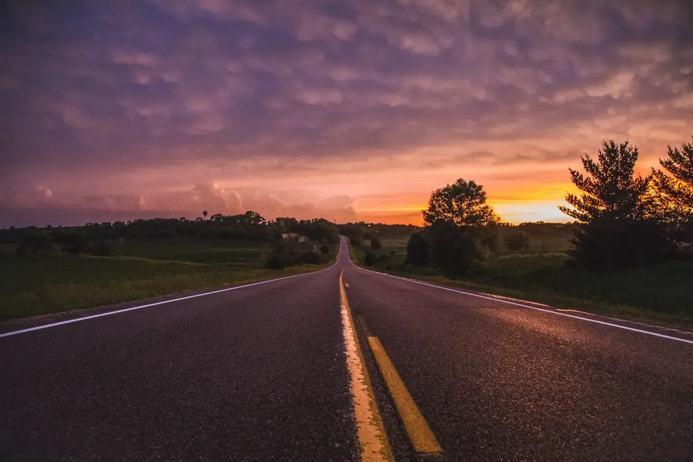 Empty road at sunset under a purple sky, with trees silhouetted on both sides.