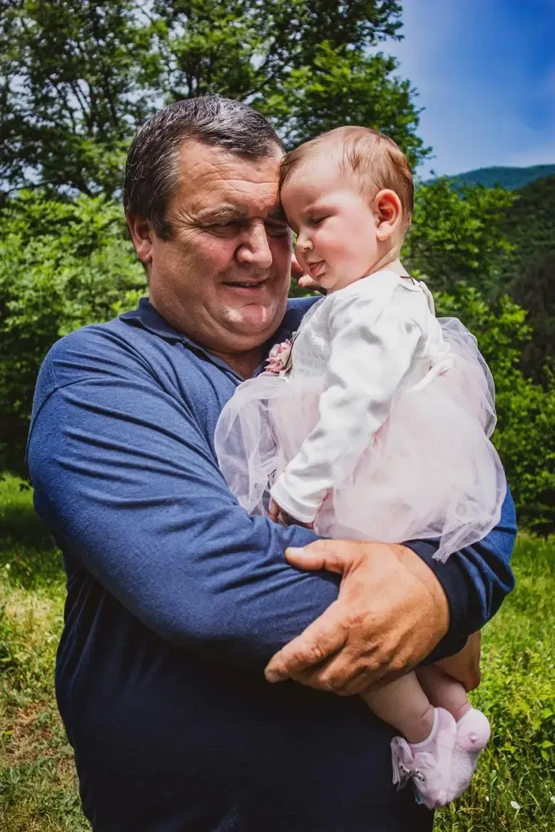 Man in blue shirt holding a baby outdoors with green trees and blue sky in the background