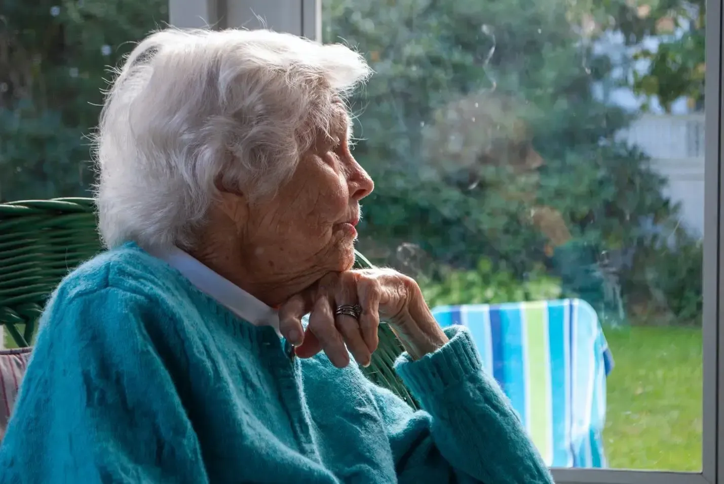 Elderly woman in a teal sweater gazes out a window, resting her chin on her hand