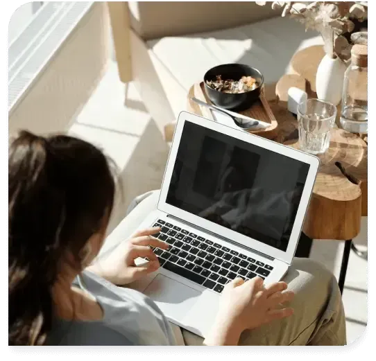 Person using a laptop at a sunlit table with coffee and a small snack nearby