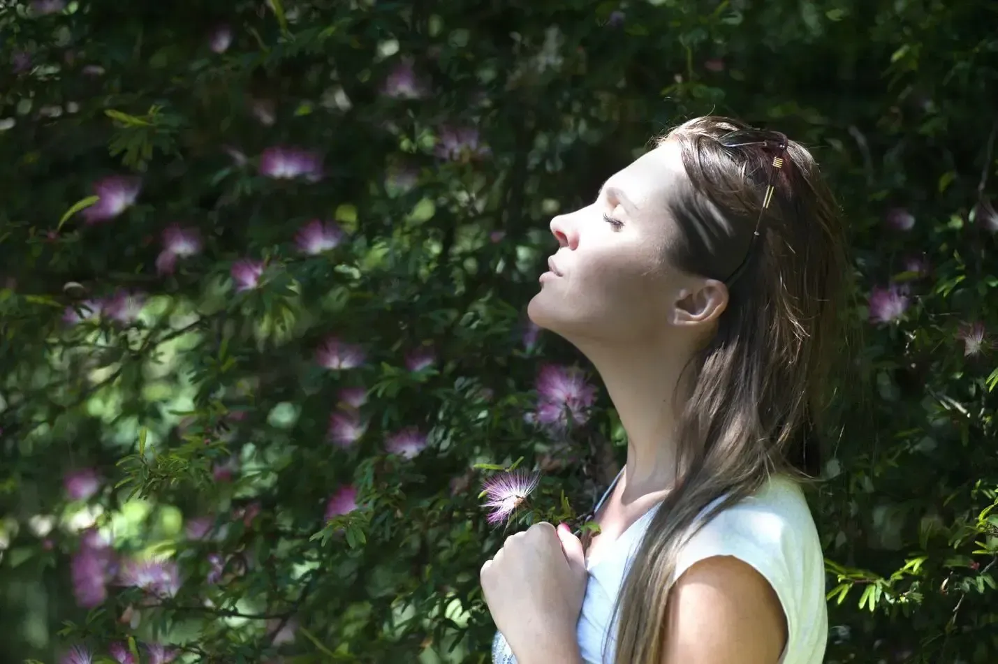 Woman in profile with eyes closed, standing by a flowering hedge in sunlight
