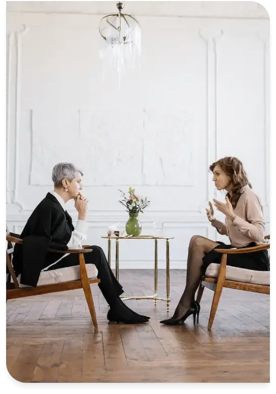 Two people seated across a small table in a bright counseling room, talking over coffee