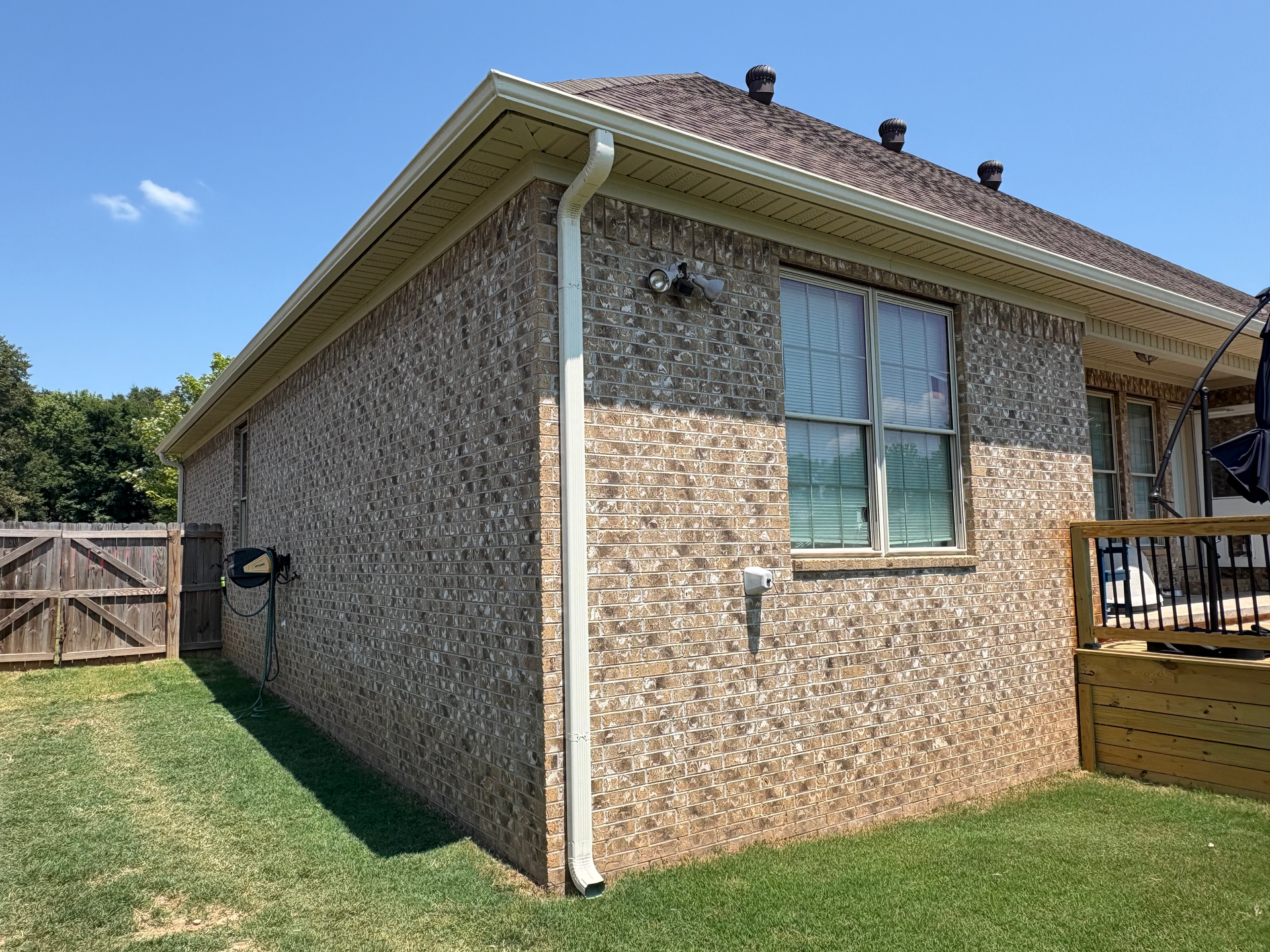 A side view of a residential brick house exterior featuring a downspout, a window, and a wooden deck under a blue sky.