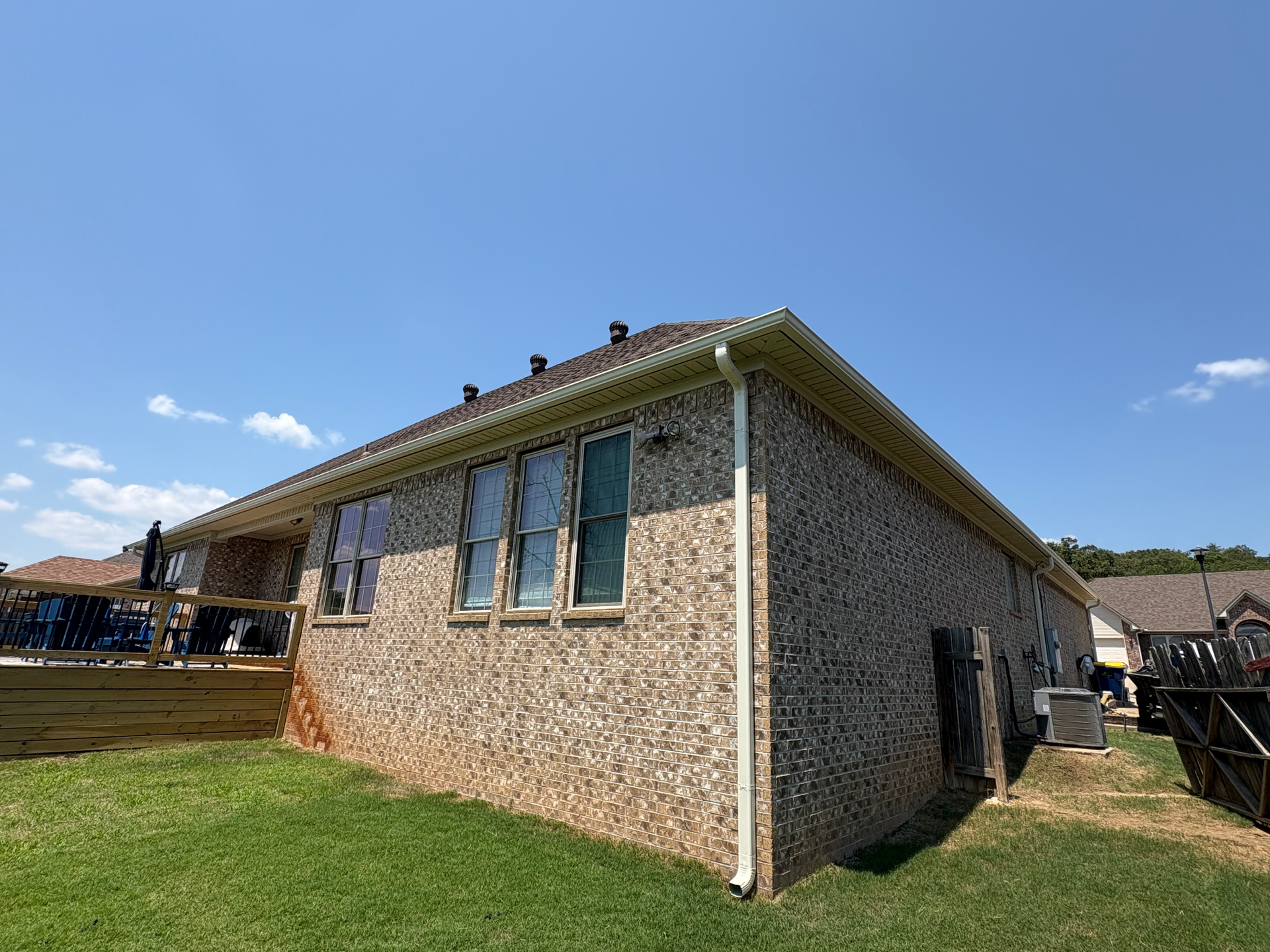 The side and rear exterior of a single-story brick house with a wooden deck under a clear blue sky.