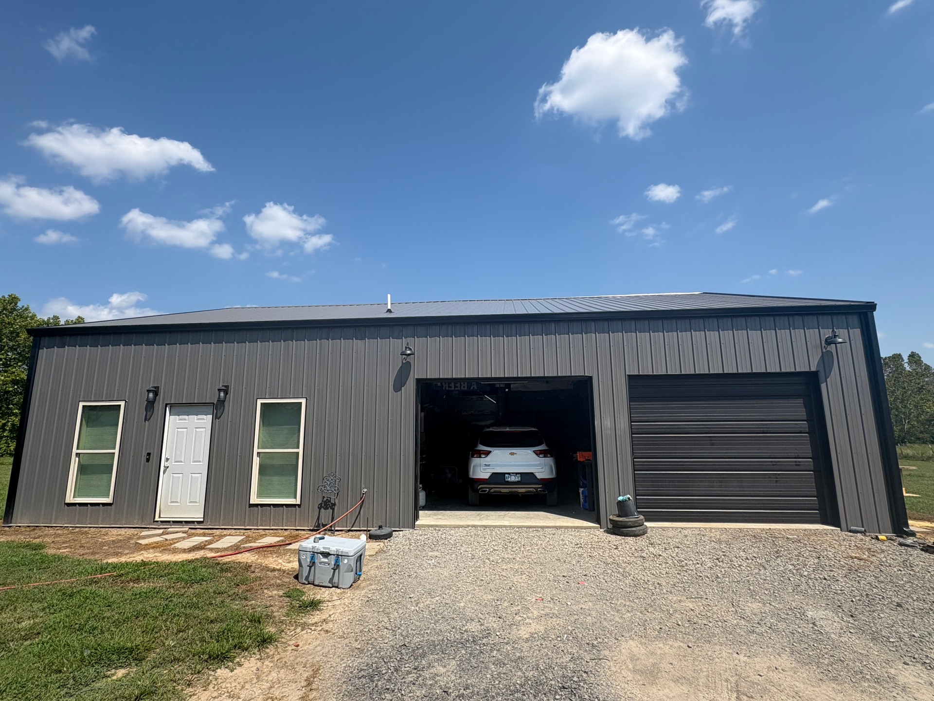 A dark gray metal workshop building with a door, two windows, and an open garage bay holding a white car under a blue sky.