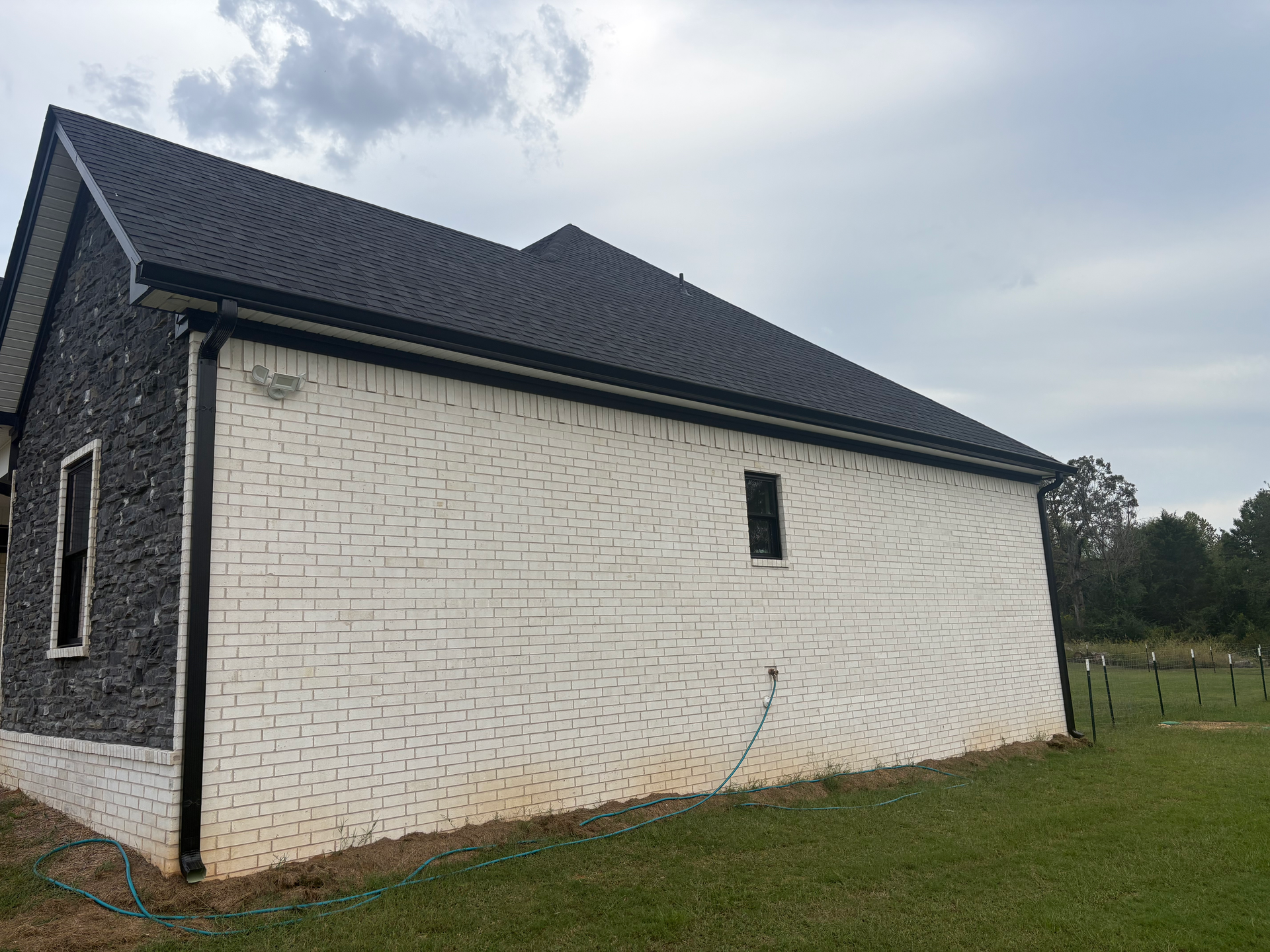 Side view of a house with white-painted brick walls, dark trim, and a shingled roof, set against a cloudy sky.