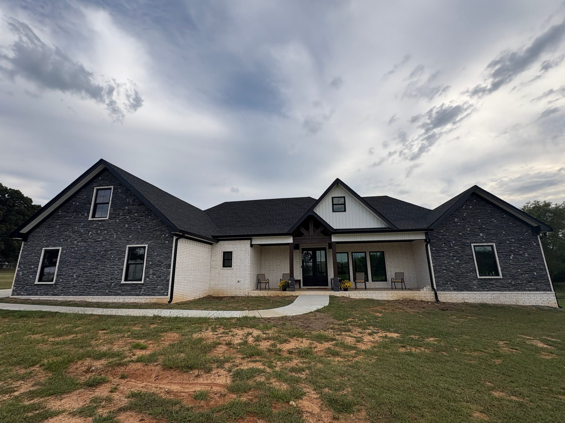 A one-story farmhouse with dark stone and white siding, a prominent covered porch, and a dark roof under a cloudy sky.