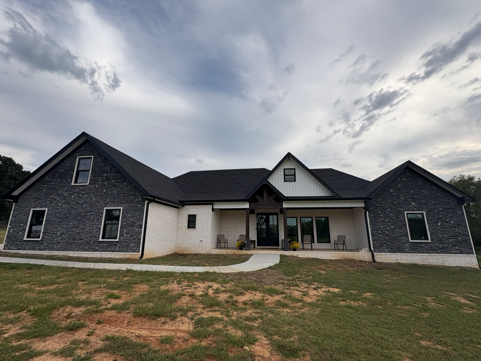 A modern farmhouse with dark stone siding, white accents, and a front porch under a cloudy sky.