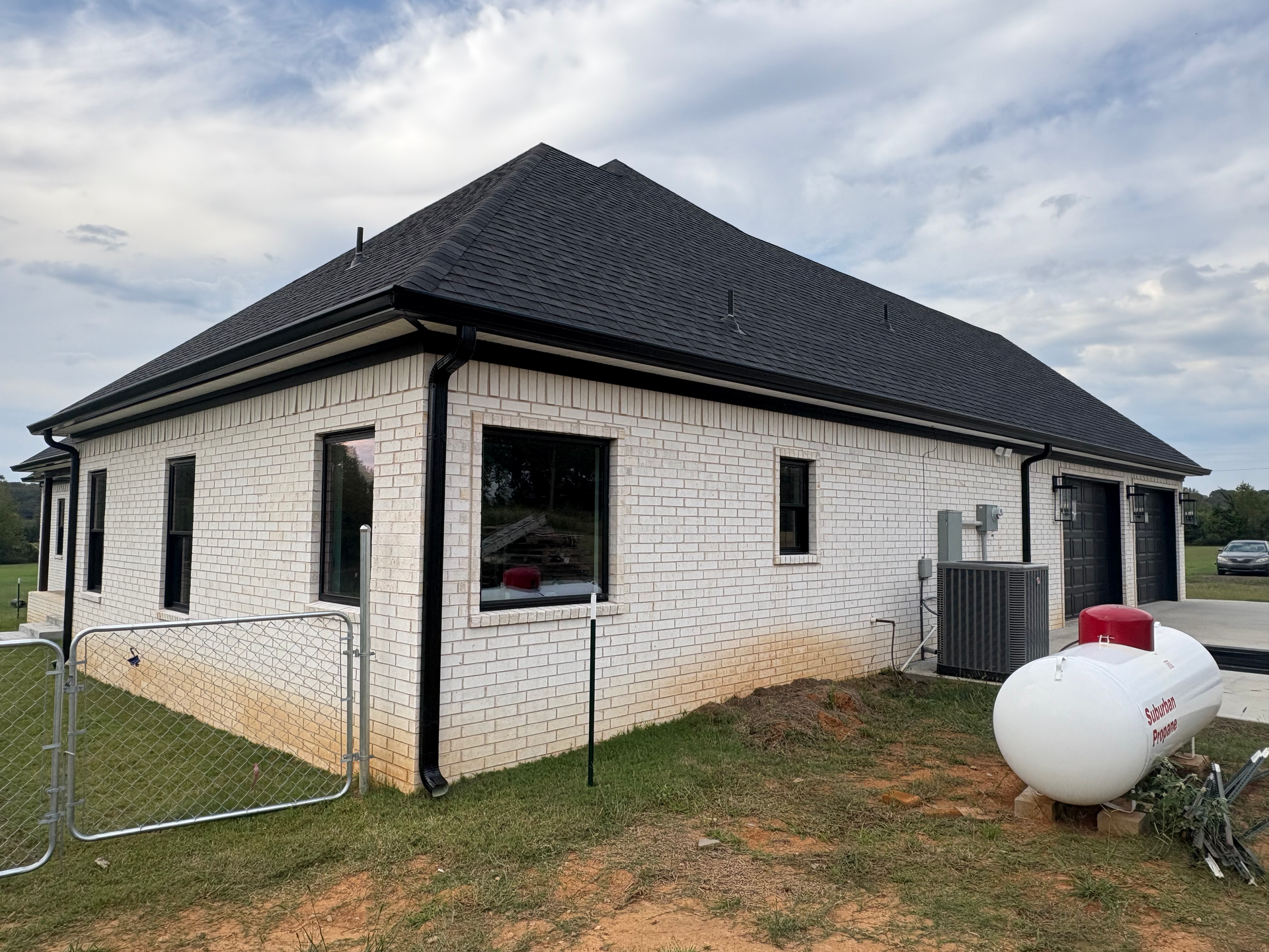 White brick house with a black roof and trim, featuring a propane tank in the yard on a cloudy day.