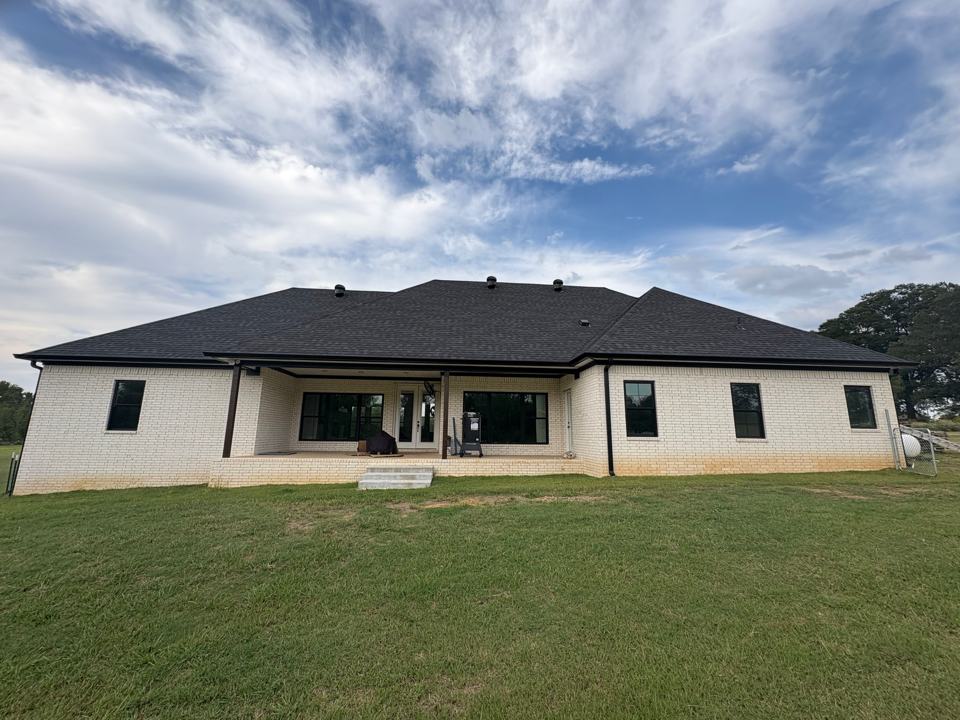 A single-story, light-colored stone house with a dark roof stands under a blue sky with scattered white clouds.