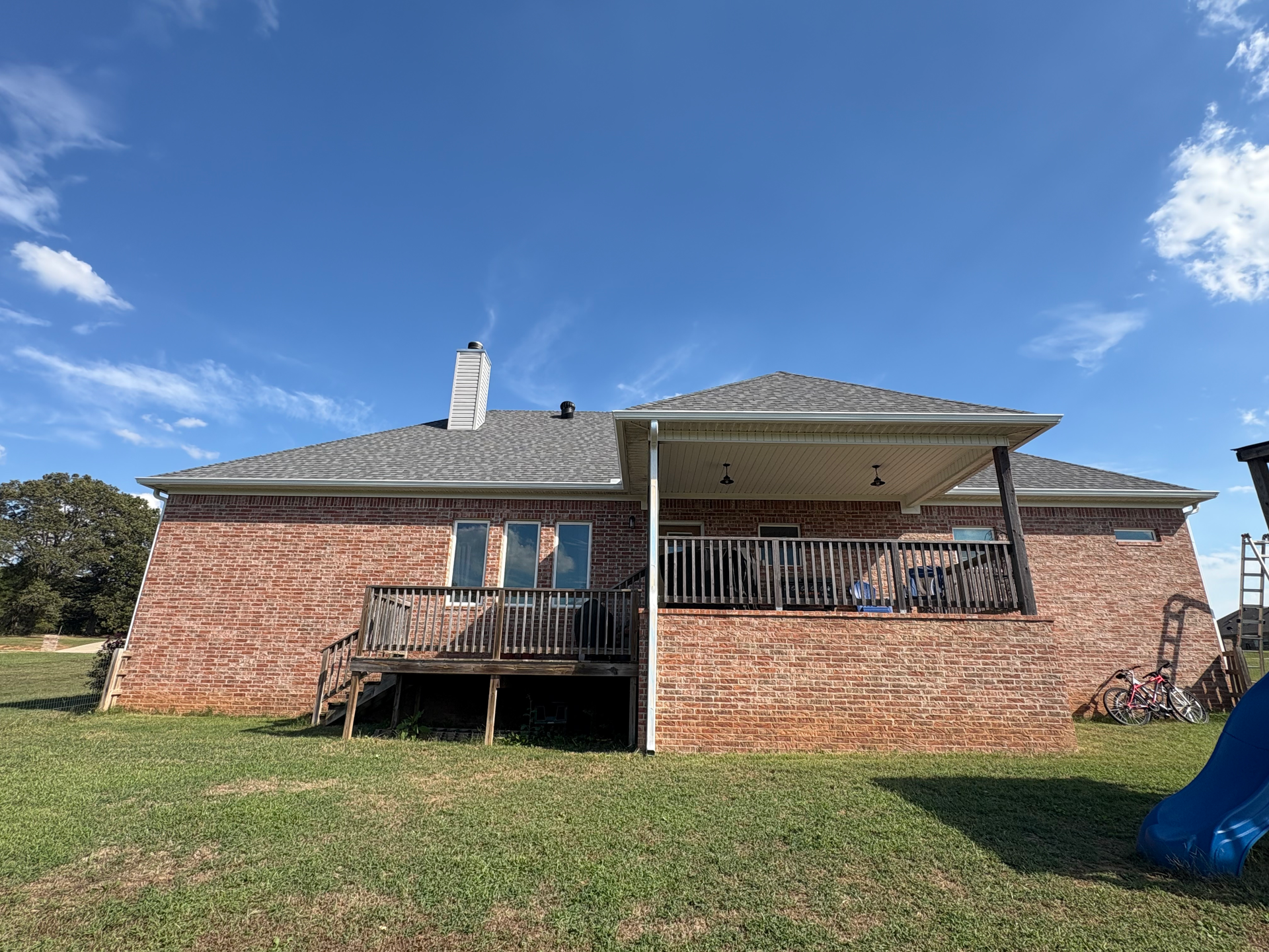 A brick house exterior with a wooden deck and a covered patio under a blue sky.