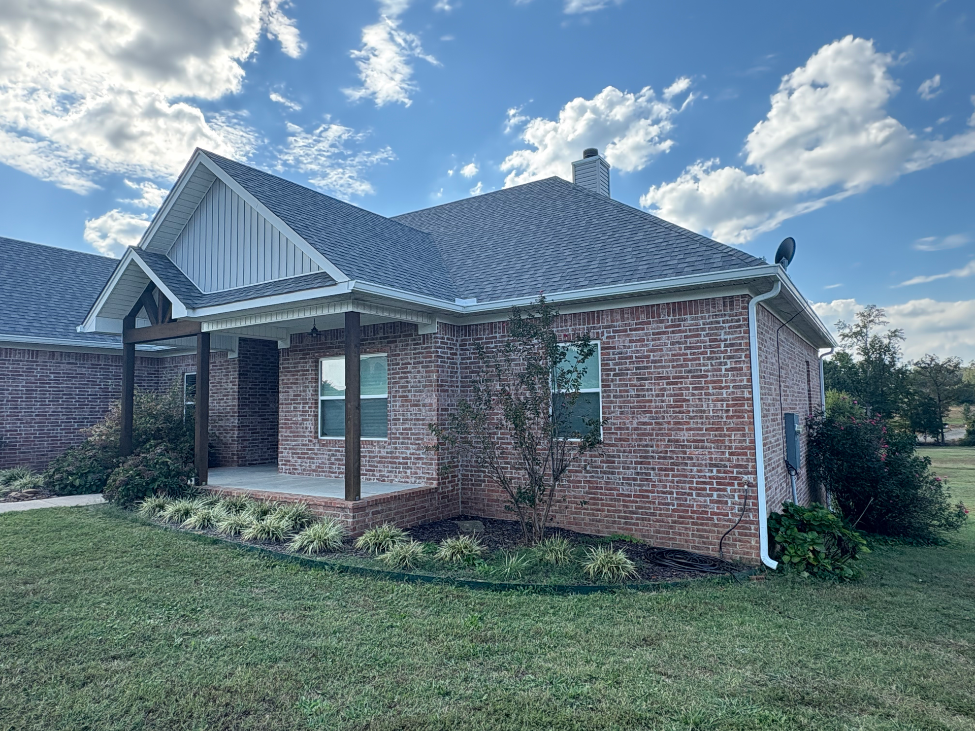 A single-story brick house with a gabled roof, covered porch, and landscaped front lawn under a bright, cloudy sky.