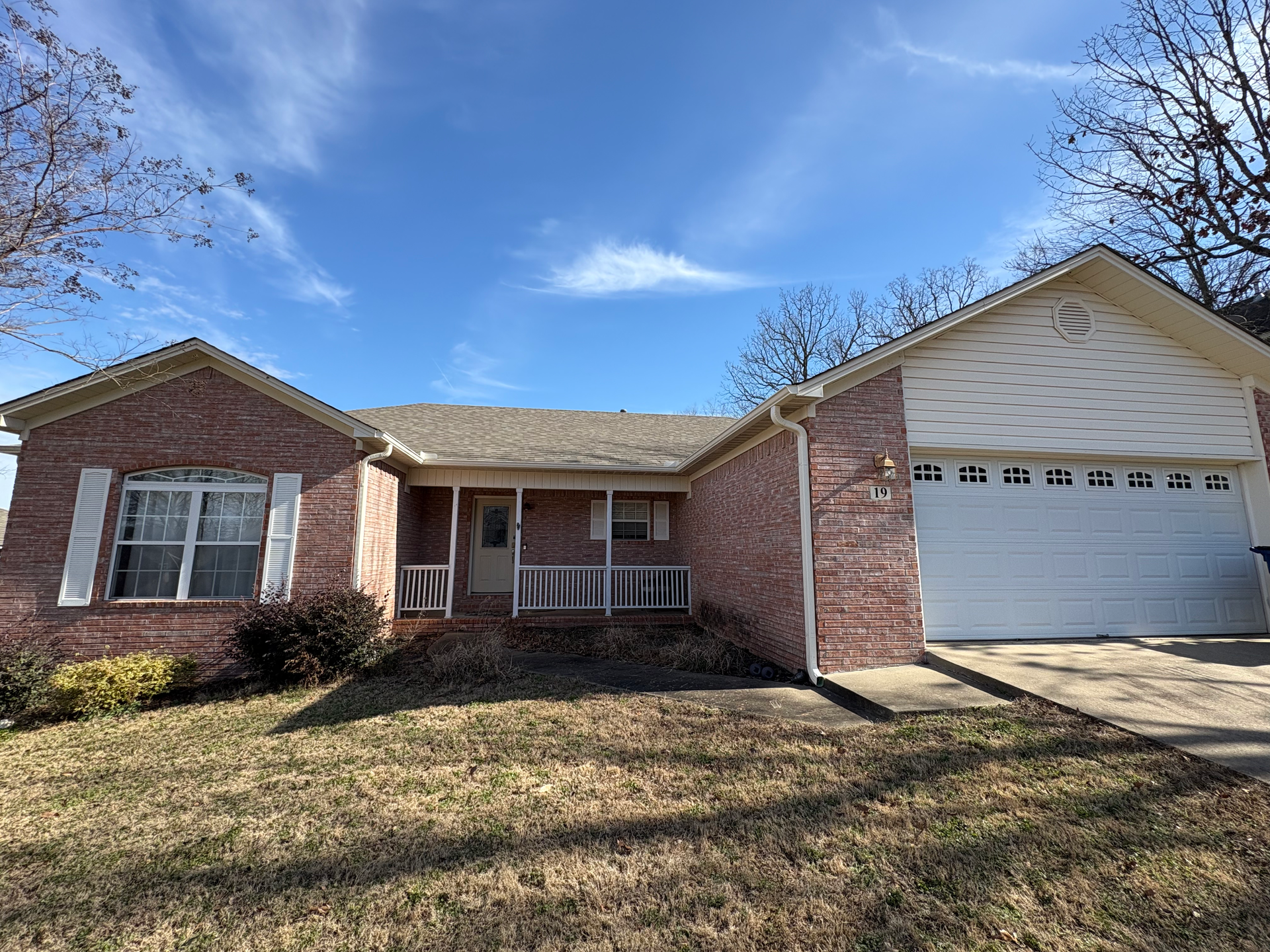 A single-story brick house with a front porch, white garage door, and light brown shingled roof under a blue sky.