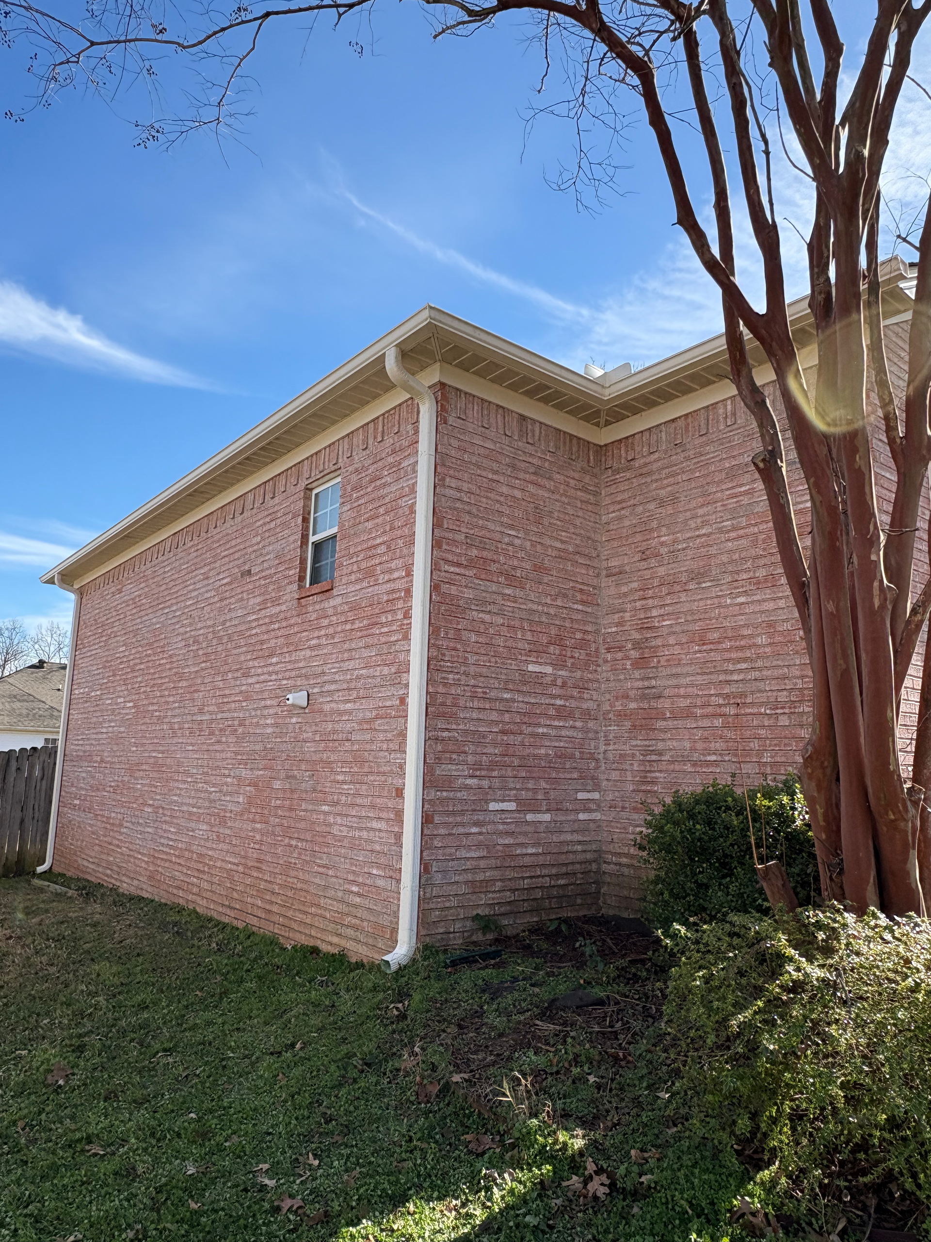 A side view of a two-story red brick house exterior under a blue sky, with a small window and a tree in the foreground.