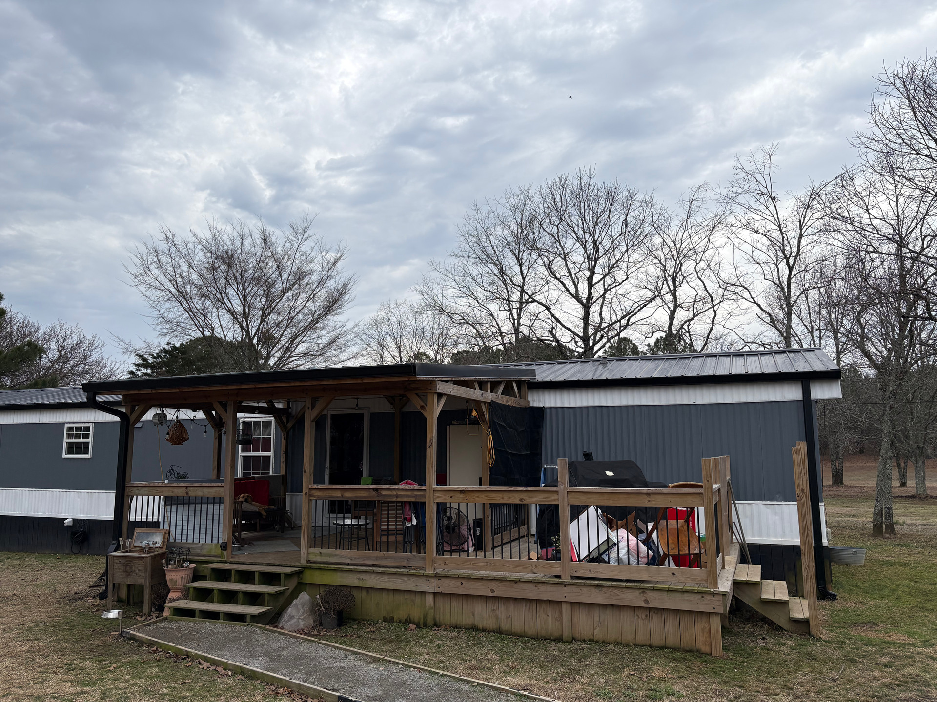 A gray mobile home with a large wooden deck and covered porch set against a cloudy sky and leafless trees.