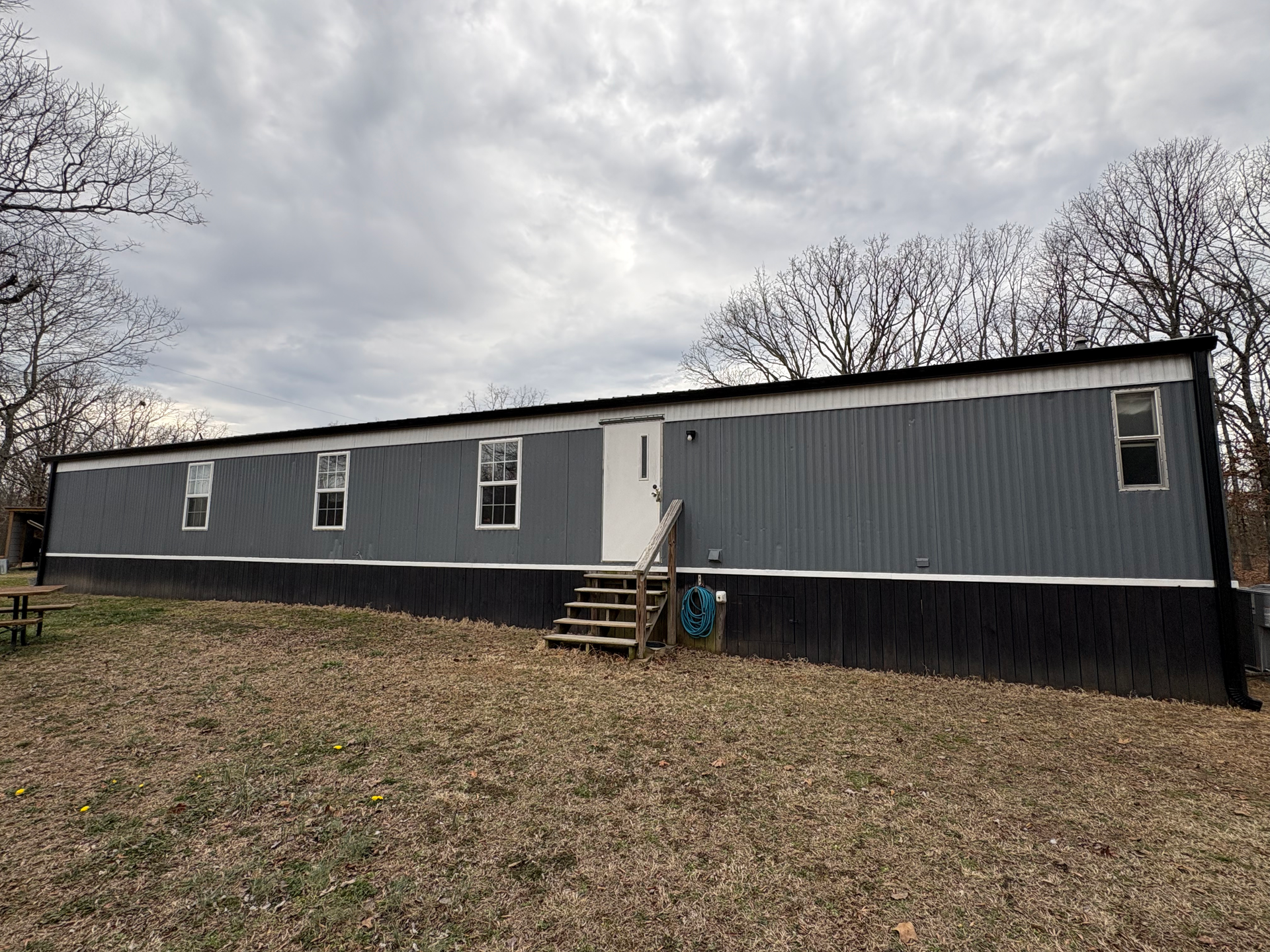 A long, gray manufactured home with a black base, white door, and wooden stairs, situated on a dry, leaf-covered lawn.