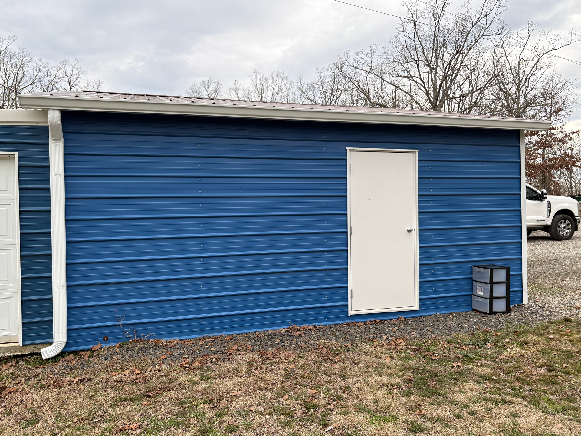 A blue corrugated metal shed with a white door and a white gutter, situated on a gravel surface outdoors.