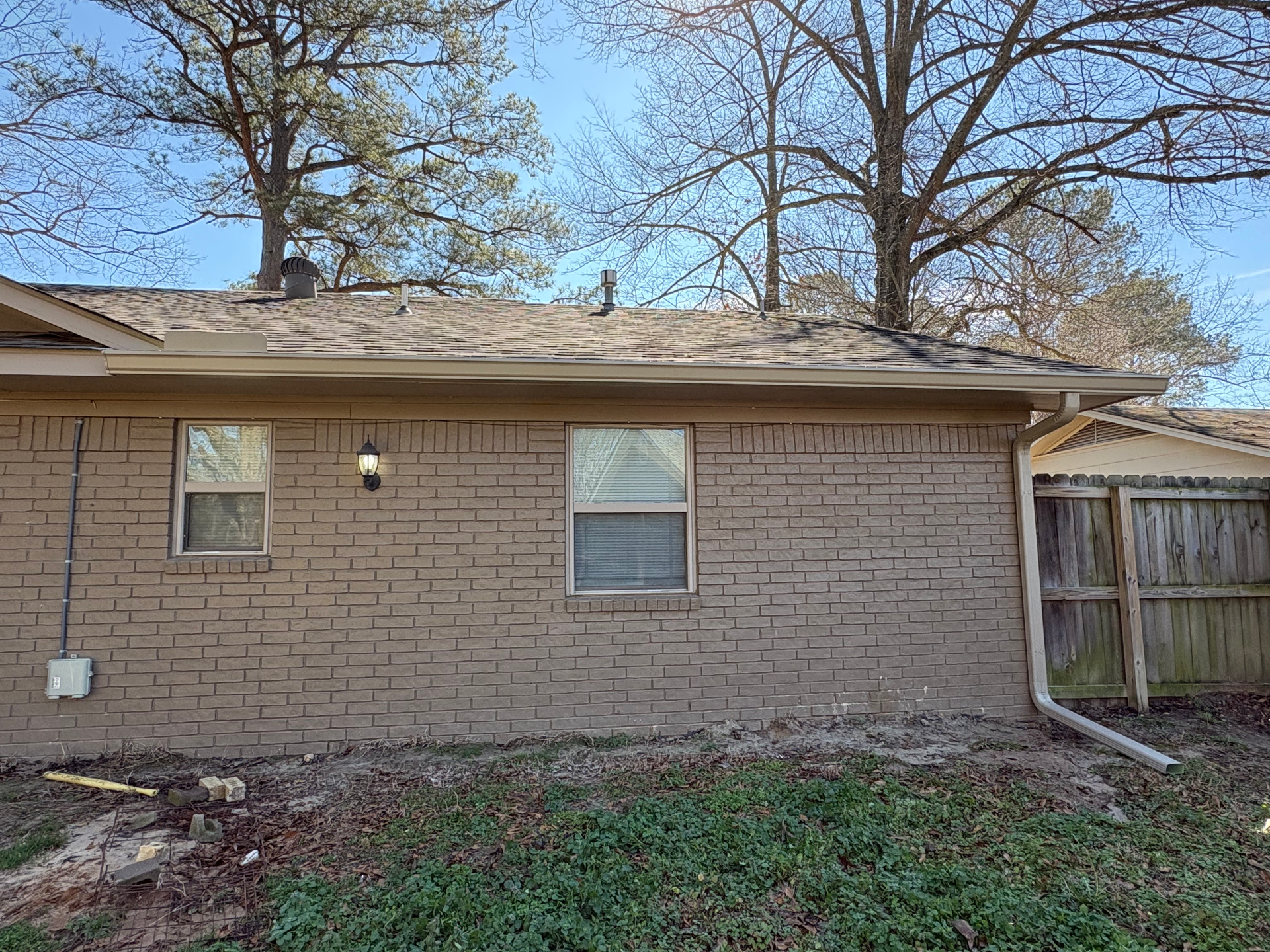 A side view of a beige brick house exterior with two windows and a gutter, situated under tall, bare trees on a sunny day.