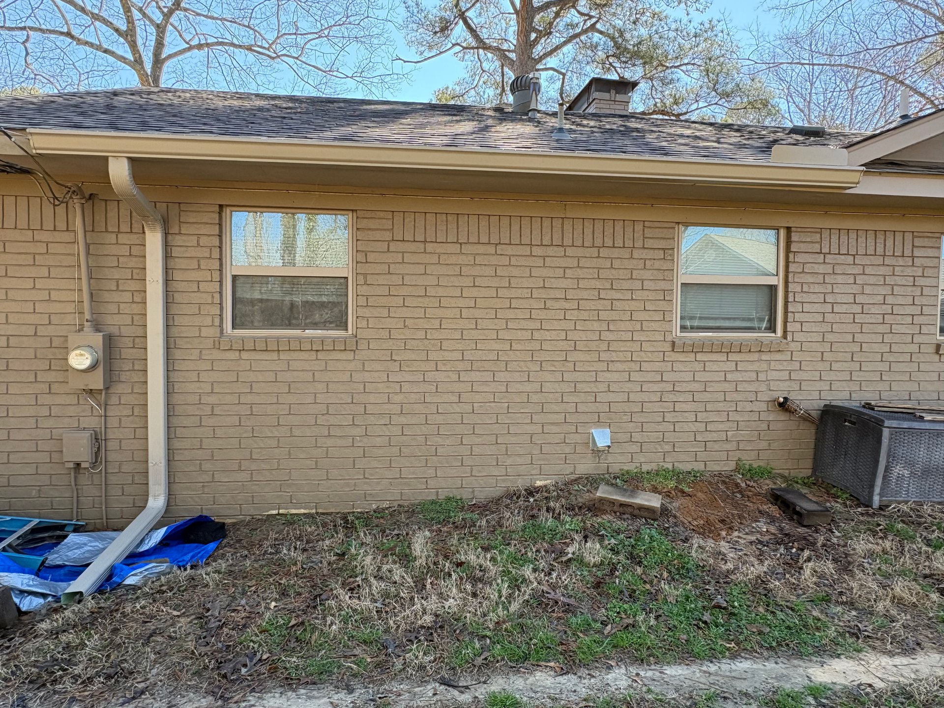 Exterior view of a tan brick house wall with two windows, a downspout, and yard debris in the foreground.
