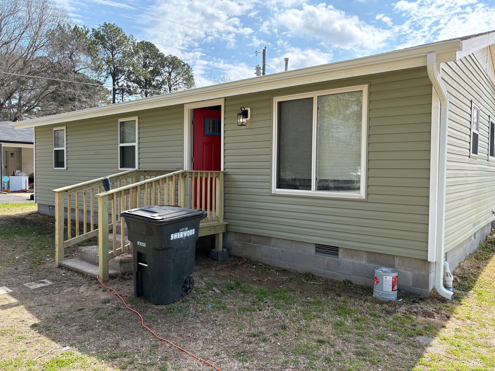 A green-sided single-story house with a red front door, a wooden wheelchair ramp, and a black trash bin in the yard.