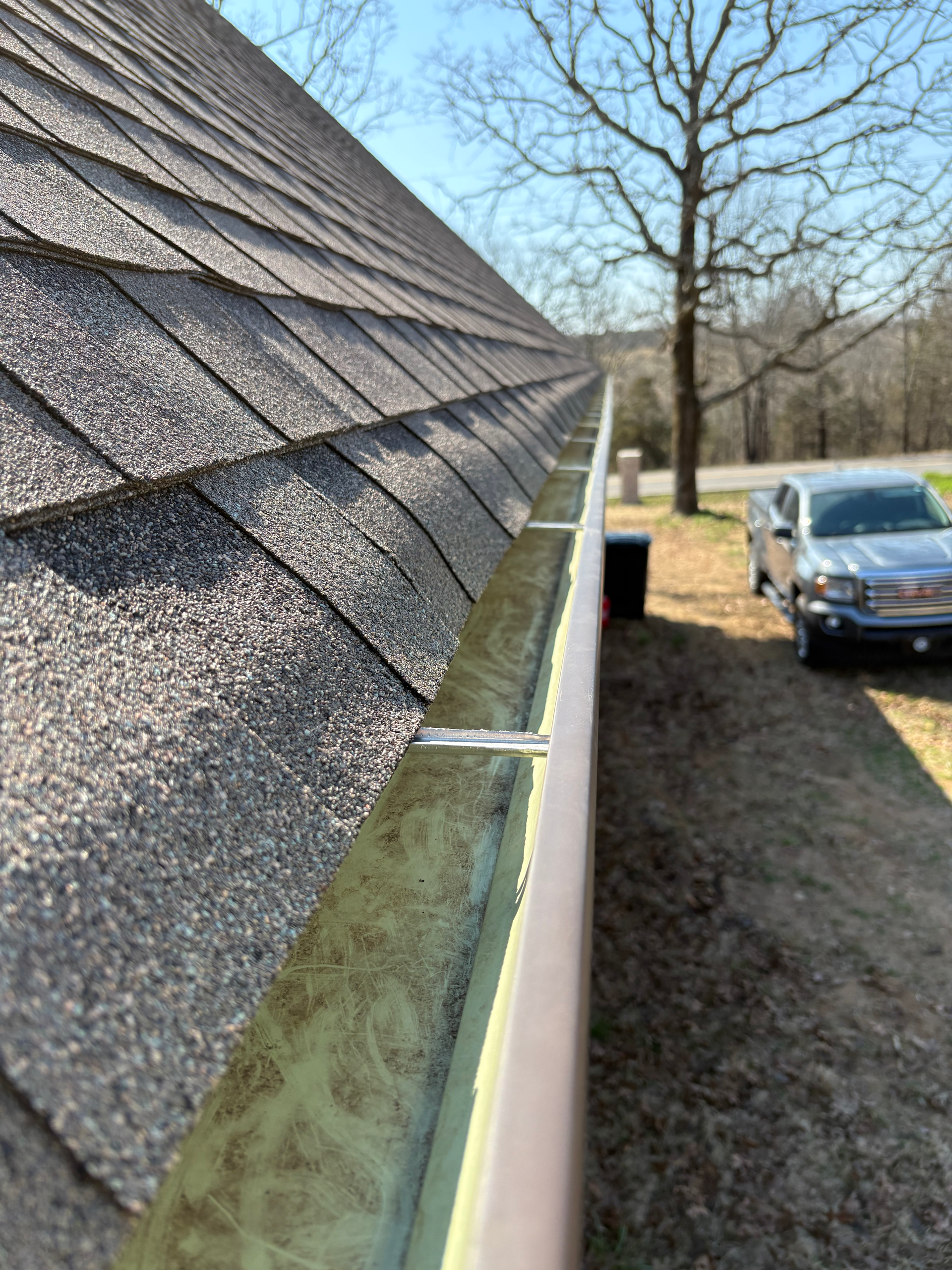 A close-up view along an exterior house gutter filled with leaves, next to a shingled roof and a truck in the distance.