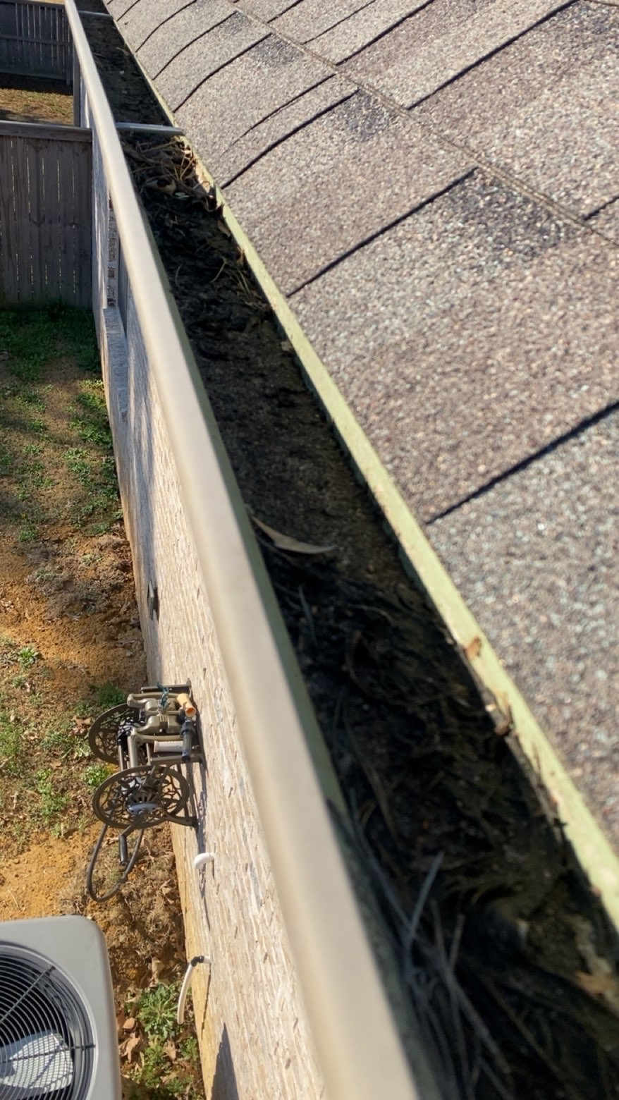 A residential rain gutter overflowing with dry, dark debris, captured from a high-angle view near a house wall.